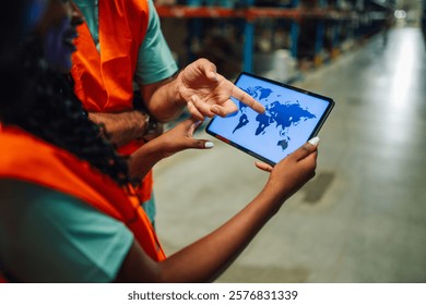 Two warehouse workers are using a digital tablet showing a world map, discussing logistics, delivery, and global shipping strategies in a modern storage facility