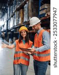 Two warehouse workers wearing safety vests and hard hats are smiling while discussing logistics and reviewing stock using a digital tablet