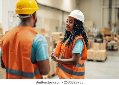 Two warehouse workers wearing safety vests and hard hats are discussing logistics inside a warehouse, with one worker holding a digital tablet - Powered by Shutterstock