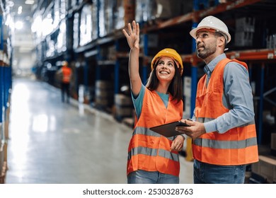 Two warehouse workers wearing safety vests are discussing logistics while using a digital tablet and pointing towards storage racks - Powered by Shutterstock