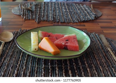 Two watermelon slices with a pineapple slice and a papaya slice on a green shiny plate with a copper spoon as a dessert after breakfast