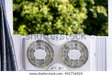 Two way window fan in home window with blurred out trees in background. 