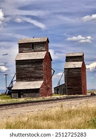Two weathered and abandoned grain elevators under a blue sky, along a train track in rural Montana.