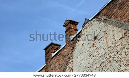 Two weathered brick chimneys extend above the roofline of an old building with crumbling plaster and exposed masonry under a light blue sky