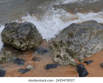 Two weathered rocks rest on a sandy beach, their surfaces worn smooth by time and tide. Gentle waves lap against the shore, leaving trails of foam in the sand. - Powered by Shutterstock