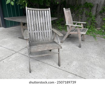 Two weathered wooden chairs are placed on a concrete patio in an outdoor garden setting. The chairs are rustic and aged, surrounded by greenery, with a wooden table in the background, creating a calm,