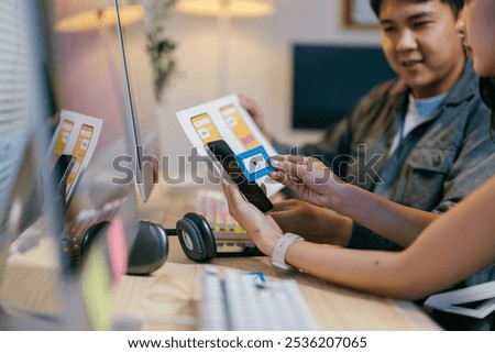 Two web designers are comparing a paper prototype with a mobile phone screen, discussing the layout and functionality of a new app