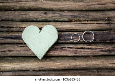 Two Wedding Rings Lie On A Wooden Table Next To A Textured Wooden Pale Green Heart