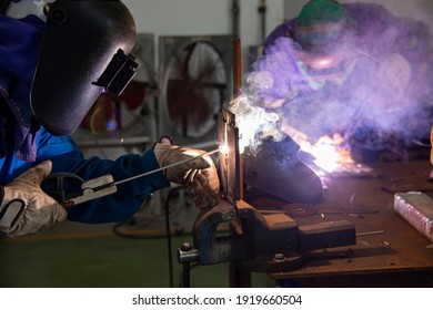 Two welder are assembling the workpiece by process shielded metal arc welding (SMAW). Welder in blue uniform, leather gloves, welding mask. The workers are testing a welding in the workshop.