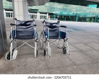 Two Wheelchairs Are Lined Up In Front Of A Hospital Or Shopping Mall Waiting To Serve The Elderly, Sick Or Handicapped. The Wheelchair Is Made Of Steel With Dark Blue Seats And Large Black Wheels.