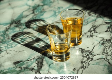 Two whiskey shots casting shadows on a marble surface in sunlight