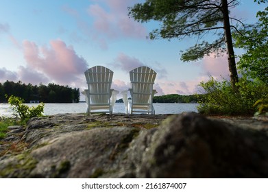 Two white Adirondack chairs on a rock formation facing the blue water of a lake in Muskoka, Ontario Canada. Beautiful sunset clouds are visible in the sky.