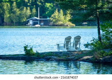 Two white Adirondack chairs on a rock formation facing the blue water of a lake in Muskoka, Ontario Canada. A cottage is visible in background.