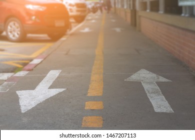 Two White Arrow Sign And Yellow Line Shown On Surface Cement Footpath And Blurred Group Car Parking On Sideroad. Blurry People Walking On Walkway With Symbol Direction And Brown Brick Wall Background.