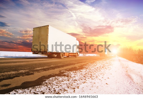 Two white box\
trucks with space for text on the countryside winter road with snow\
against a sky with a sunset