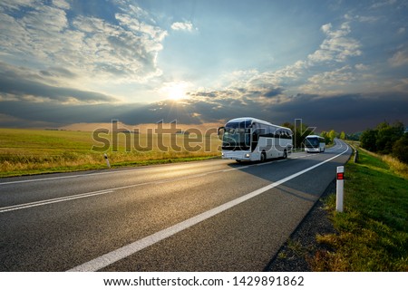 Two white buses traveling on the asphalt road in rural landscape at sunset with dramatic clouds                               