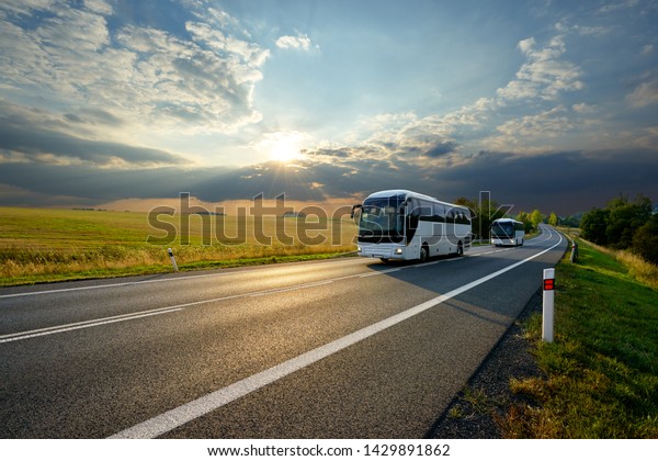 Two white buses traveling on the asphalt road in rural\
landscape at sunset with dramatic clouds                           \
   