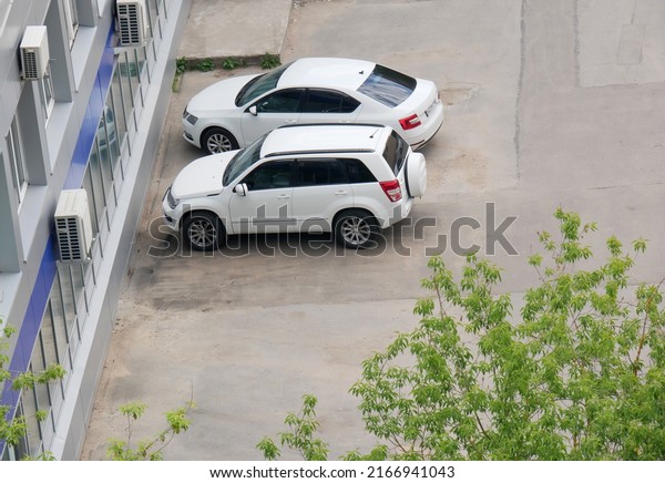 Two white cars on asphalt lot in city.  Spontaneous\
parking of transport. Сoncept of undeveloped infrastructure.\
Reflections in rear window of cars and building windows.Copy space.\
Top view.  
