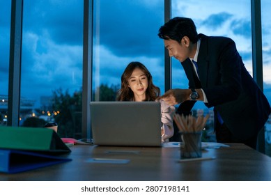 Two white collar workers sit close together at a desk, focused on a laptop screen, working late into the night in an illuminated office.
