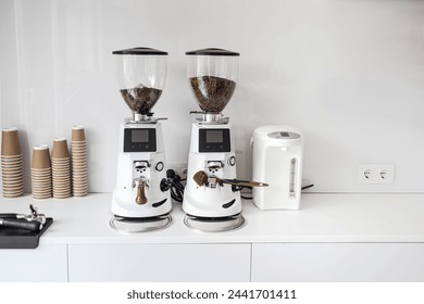 Two white commercial coffee grinders with digital displays, ready for use in a cafe setting, flanked by stacked paper cups and a white electric kettle. - Powered by Shutterstock