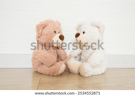 Two white and light brown soft teddy bears looking at each other and sitting on wooden floor at light gray wall background. Closeup. Children communication concept. Front view.