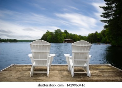 Two white Muskoka chairs on a dock overlooking a lake in Ontario Canada. Across the calm water there's a brown cottage nestled between green trees.