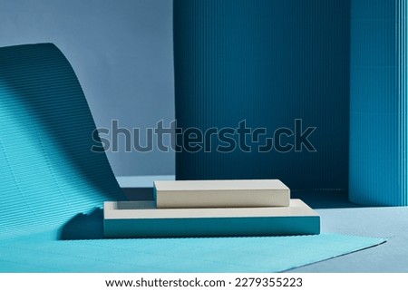 Two white rectangular empty podiums stacked on dark background. Blue folds form the soft undulating wall and rug. Minimal concept for product presentation