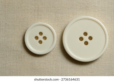 Two white sewing buttons of different sizes on natural linen fabric representing choice, comparison, tailoring, clothing design and craftsmanship. Minimal sewing concept with copy space for fashion, handmade, textile industry, apparel manufacturing and creative design.