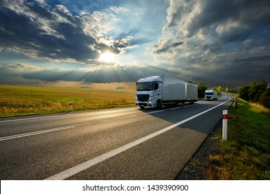 Two white trucks driving on the asphalt road in rural landscape at sunset with dramatic clouds                               