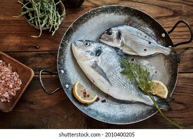 Two whole fresh raw gilt-head bream in a vintage metal wok or skillet with pink Himalayan salt, lemon and herbs for seasoning in a top down view on a rustic table - Powered by Shutterstock