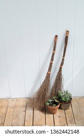 Two Wicker Broomsticks Resting Against A White Wall With A Potted English Ivy And A Potted Lavender Plant On A Worn Wooden Floor.