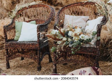 Two wicker chairs with white cushions and a bouquet of flowers on it. Rattan chairs with straw bales in the background. Boho style