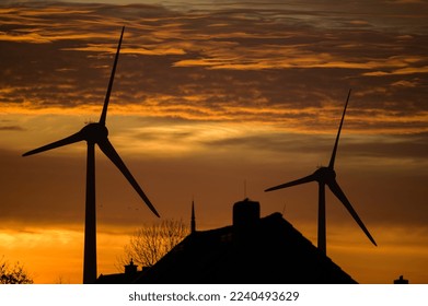 Two wind turbines set against spectacular orange sunrise and clouds in the sky next to a house with birds passing by in the distance - Powered by Shutterstock