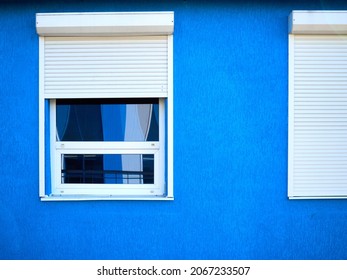 two windows of a part of the building, white shutters, blinds are closed at one window, there is a reflection of the street in the other window, the walls of the building are painted blue