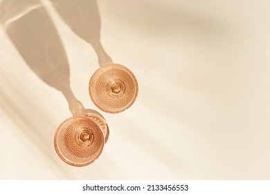 Two Wine Glasses From Pale Pink Color Glass With Palm Leaf Shadows From Sun, Trendy Goblets Style Wine Glasses With White Wine On Beige Background. Pastel Colors, Monochrome Image, Top View Glassware