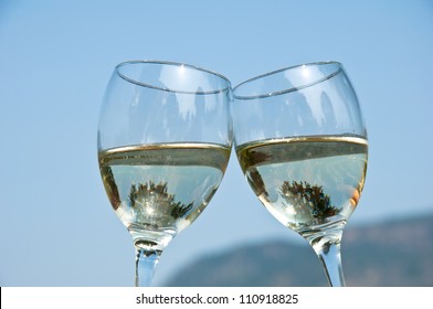 Two Wineglasses Filled With White Wine Clinking Each Other Outside On A Summer Day.