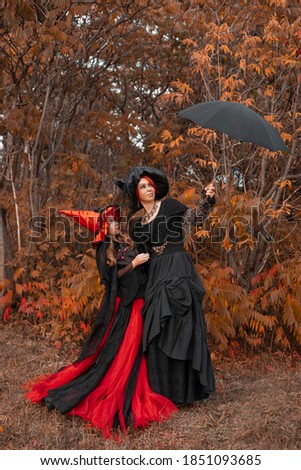 two witches, mother and daughter, in the black and red dresses and pointed hats standing in an autumn forest under the umbrella, halloween costumes