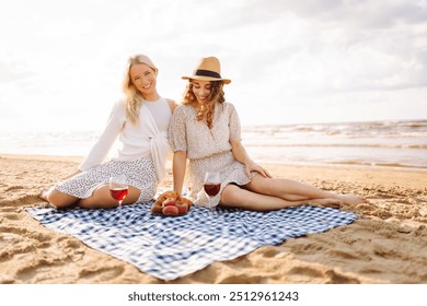 Two woman friends enjoying red wine on a sandy beach during sunset, capturing a joyful moment filled with laughter and warmth.Hen-party. - Powered by Shutterstock