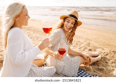 Two woman friends enjoying red wine on a sandy beach during sunset, capturing a joyful moment filled with laughter and warmth.Hen-party. - Powered by Shutterstock