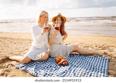 Two woman friends enjoying red wine on a sandy beach during sunset, capturing a joyful moment filled with laughter and warmth.Hen-party. - Powered by Shutterstock