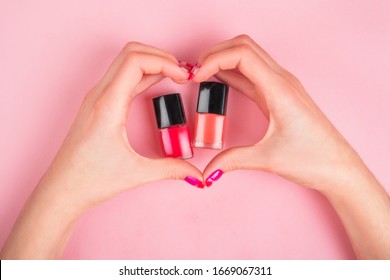 Two woman hands and bright manicure making heart shape to nail polish bottles isolated pink background 