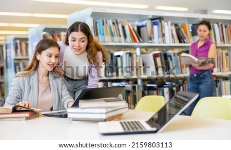 Two woman students, who are preparing a report on a laptop in the university library, discuss studying issues