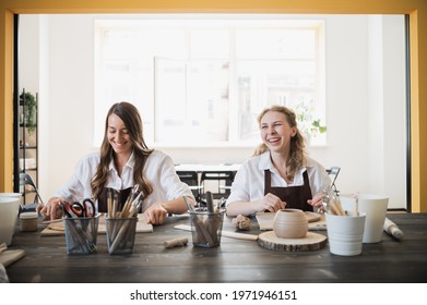 Two women in aprons sitting by table, kneading clay and making earthenware at lesson - Shutterstock ID 1971946151