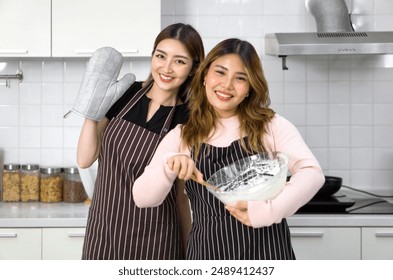 Two women in aprons smiling in the kitchen, one holding an oven mitt, the other mixing batter in a bowl. - Powered by Shutterstock