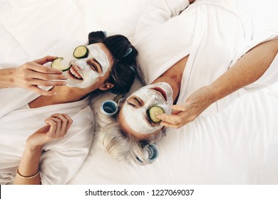 Two Women In Bathrobes Lying On Bed Wearing Beauty Face Pack With Cucumber Slices. Mother And Daughter Relaxing On Bed Wearing Face Pack And Roller Curls On Hair.