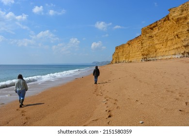 Two women from behind walking on the seashore beneath cliffs on Hive Beach, Dorset, England