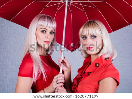 Two women with blond hair stand together under a large umbrella.