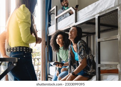 Two women, Caucasian and Brazilian, laughing with a smartphone, surrounded by friends in a hostel room, boy on upper bunk laughs along - Powered by Shutterstock