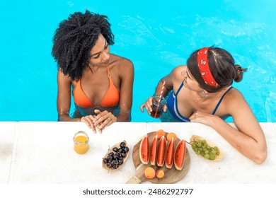 Two women chatting by the pool - enjoying summer fun - fruit and drinks, friends relaxing - swimwear and healthy snacks, outdoor poolside - casual conversation. - Powered by Shutterstock