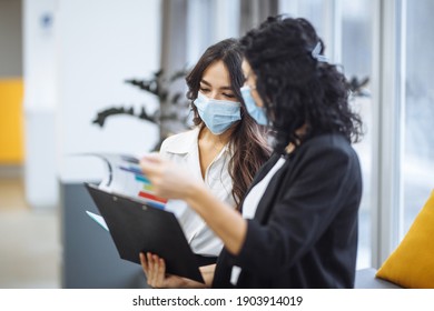 Two women checking charts and discussing business trends in the office. Females colleagues wearing medical masks sit on the couch at a working place. Coronavirus prevention. Colaboration concept